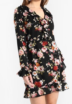 Femme portant une robe noire à fleurs avec un col volanté, des manches longues et un ourlet avec des fleurs roses, rouges et blanches accompagnées de feuilles vertes.
