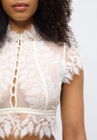 Δαντελένιο cropped τοπ με κοντά μανίκια, με σχέδια λουλουδιών, ψηλό γιακά και λεπτομέρειες με κουμπιά. Απαλή, διάφανη υφή σε κρεμ χρώμα.