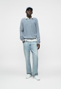 Gaiši zils ribots adīts džemperis virs balta kreklveida krekla, kombinēts ar gaiši ziliem taisnām kājām džinsiem un baltām sporta kedām.