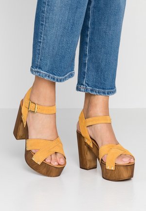 Pies que llevan sandalias de plataforma de ante amarillas con tacones de bloque de madera y correas al tobillo, combinadas con jeans de mezclilla azul recortados.