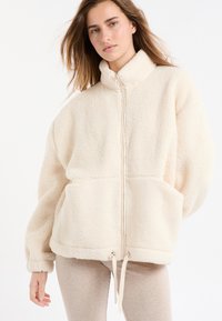 Kremowa kurtka z polaru z wysokim kołnierzem, zamkiem błyskawicznym z przodu, elastycznymi mankietami i dwoma bocznymi kieszeniami. Posiada regulowane sznurki u dołu.