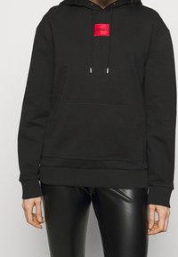 Sudadera negra con bolsillo delantero, cordón, parche de logo rojo y puños y dobladillo acanalados. Llevada con leggings negros brillantes.
