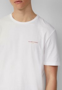 Witte katoenen t-shirt met een ronde hals en korte mouwen, voorzien van afgedrukte coördinaten en tekst in roze en beige aan de linkerkant.