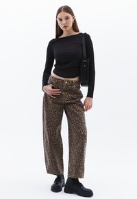 Schwarzes langärmliges Crop-Top und braune weit geschnittene Hose mit Leopardenmuster. Das Model trägt schwarze Schnürstiefel und eine schwarze Umhängetasche.