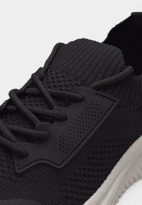 Μαύρο πλεκτό αθλητικό παπούτσι με υφασμένο επάνω μέρος, χοντρά κορδόνια και λευκή σόλα, εμφανίζοντας λεπτομέρειες από μπροστά και από το πλάι.