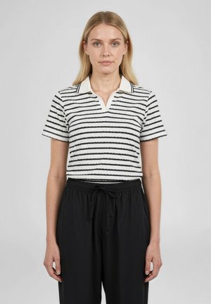 Frau mit blondem Haar, die ein weißes und schwarzes, kurzärmeliges Polohemd mit Streifen und schwarze, mit Kordelzug versehene Hosen trägt, steht vor einem schlichten Hintergrund.