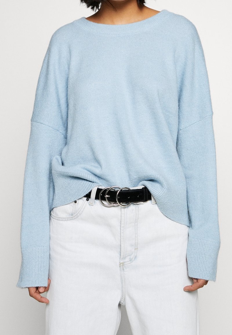 Jasnoniebieski sweter z dzianiny o luźnym kroju, opadające ramiona, ściągacze na mankietach, noszony na białych dżinsach z wysokim stanem, w połączeniu z czarnym paskiem.