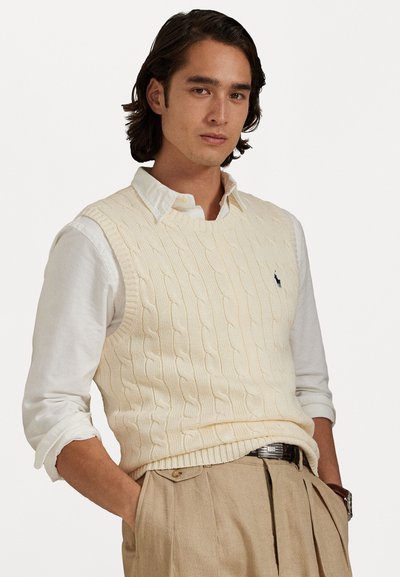 Chaleco de punto trenzado crema, superpuesto a una camisa blanca con cuello, con dobladillo acanalado y sutil detalle del logo. Combinado con pantalones beige.