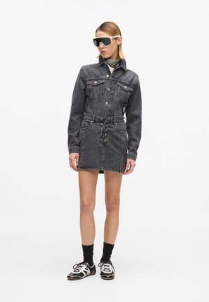 Γυναίκα που φοράει σκούρο γκρι τζιν μπουφάν και ταιριαστή φούστα με κορδόνια, μαύρες κάλτσες, ασπρόμαυρα παπούτσια και υπερμεγέθη λευκά γυαλιά ηλίου.