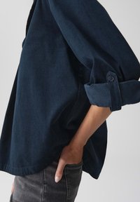 Persona che indossa una camicia di velluto a coste blu scuro con le maniche arrotolate e jeans grigi, con una mano casualmente infilata in tasca.