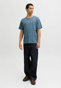 Camiseta para hombre de color verde azulado con un pequeño logo, combinada con vaqueros anchos y oscuros. El conjunto presenta un ajuste relajado y una textura suave. Zapatos marrones.