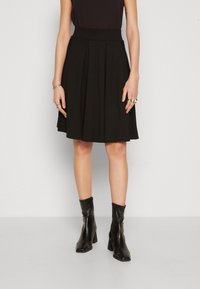 Černá skládací sukně s vysokým pasem, vyrobená z měkké tkaniny; zkombinovaná s černými kotníkovými botami se čtveratou špičkou a blokovým podpatkem.