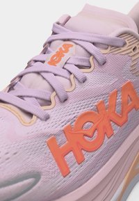 Ανοιχτό μωβ παπούτσι για τρέξιμο Hoka με πορτοκαλί λογότυπο HOKA, μωβ κορδόνια και μπεζ λεπτομέρειες στο πλεκτό πάνω μέρος και μαξιλαρωμένη γλώσσα.