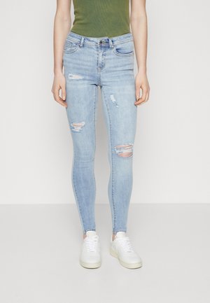 Personne portant un jean skinny bleu clair déchiré, des baskets blanches et un haut sans manches vert olive, debout devant un fond uni.