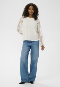 Haut blanc à manches longues en dentelle fleurie, associé à un jean bleu clair à jambes larges et des talons noirs pointus. Tissu lisse avec une coupe décontractée.