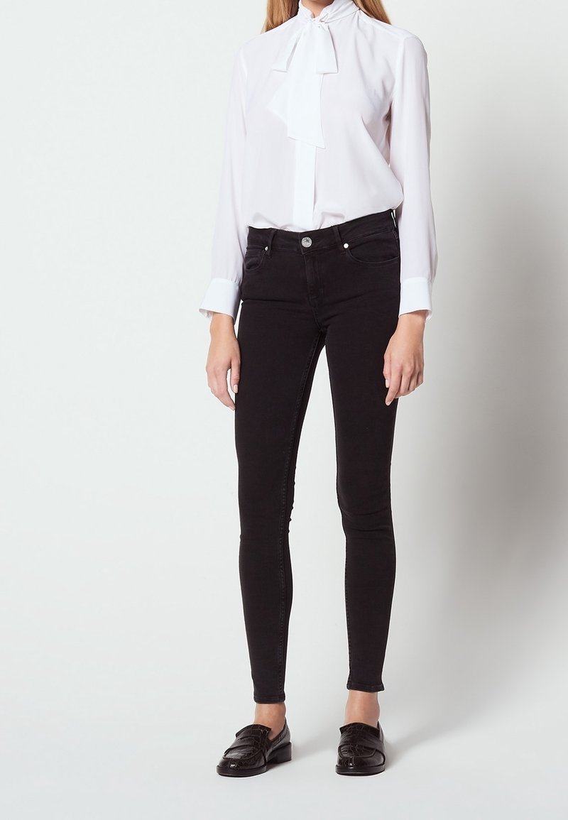 Jeans escoceses negros ajustados de mezclilla con un ligero estiramiento, combinados con una blusa blanca con un detalle de lazo en el collar y mangas largas.