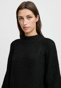 Chunky zwarte gebreide trui met een hoge halslijn, geribbelde textuur en een relaxte pasvorm. Kenmerken zijn ingeklede schouders en een zachte, comfortabele uitstraling.