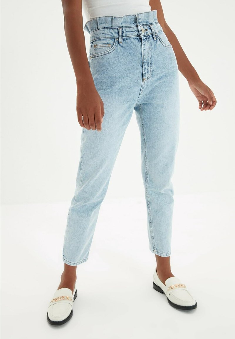 Personne portant un jean taille haute bleu clair et des mocassins blancs avec un détail en chaîne dorée, se tenant devant un fond blanc uni.