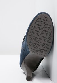 Zapato de tacón alto azul marino con acabado de ante, que presenta una suela negra texturizada embosada con el nombre de la marca "Tamaris".