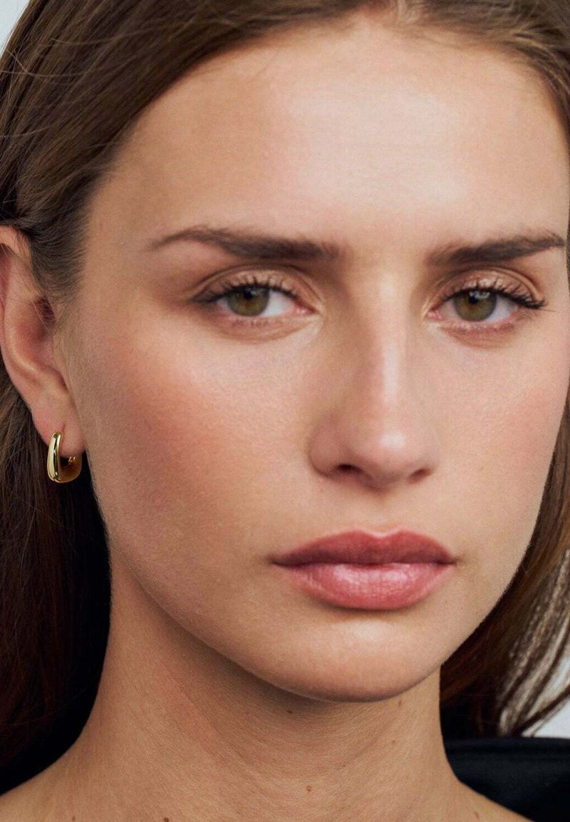 Närbild av ett ansikte med brunt hår, olivfärgad hudton och naturlig sminkning; har guldörhängen och fylliga läppar.