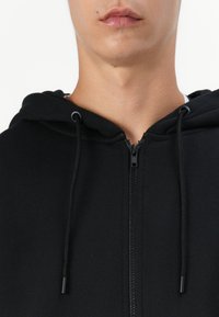Sudadera negra con cremallera frontal, cordones y una textura suave. Presenta un diseño sencillo, que muestra un estilo limpio y minimalista.