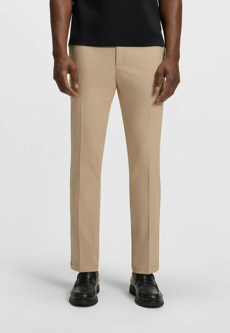 Beżowe spodnie w kant o gładkiej fakturze i prostym kroju, z subtelnym wzorem splotu, zestawione z czarnymi loafersami.