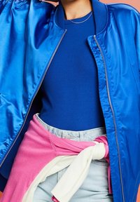 Personne portant une veste bomber bleu brillant sur une chemise bleue, avec un pull rose et blanc attaché autour d'un pantalon en jean clair.