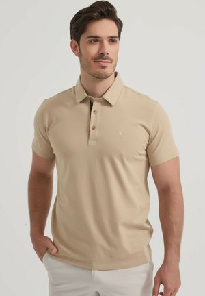 Homme portant un polo beige à manches courtes et un pantalon blanc, une main dans la poche, regardant légèrement sur le côté.