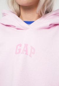 Sudadera rosa claro hecha de una tela suave, con un bolsillo canguro en la parte delantera. Presenta un pequeño logo "GAP" desvanecido en un tono rosa a juego.