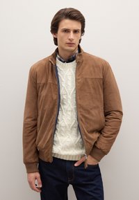 Braune Wildleder-Bomberjacke mit Reißverschluss vorne, gerippten Bündchen und flachem Kragen, kombiniert mit einem cremefarbenen Strickpullover mit Zopfmuster und dunklen Jeans.