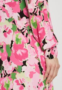 Gėlių suknelė, kuri pasižymi ryškiu rožiniu fonu su žaliais ir juodais akcentais. Lygi tekstūra su mygtukais detalėmis ant rankovių.