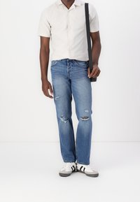 Muž v béžové košili s krátkým rukávem, roztrhaných modrých džínech, bílých teniskách s černými pruhy a s černou taškou přes rameno.