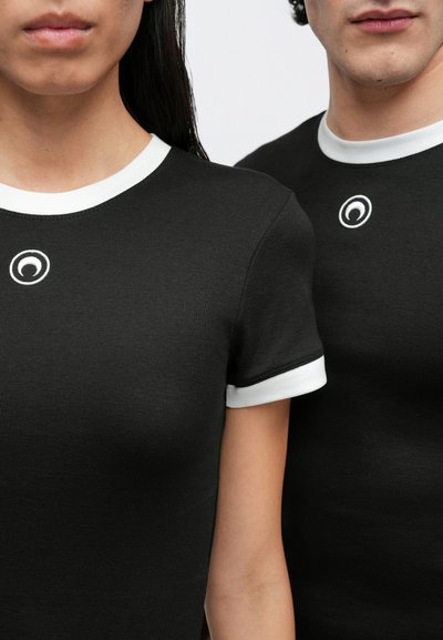 Juodos spalvos marškinėliai su apvalia iškirpte ir baltais gofruotais rankogaliais. Medvilnės audinys su apvaliu logotipu ant krūtinės. Artimas dviejų dėvėtojų vaizdas.