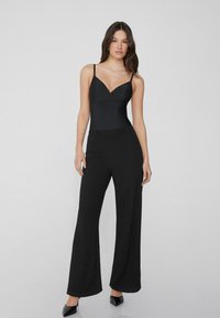 Bodysuit negro con tirantes finos y escote en V, combinado con pantalones anchos negros. Textura suave y ajuste a medida, con un diseño formal.