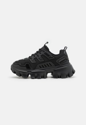 Sneaker nera chunky con suola spessa e robusta, chiusura con lacci e parte superiore in sintetico testurizzato su uno sfondo bianco neutro.