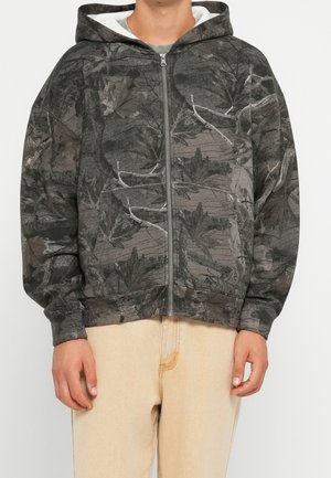 Homme portant un sweat à capuche zippé camouflage gris et noir avec poches avant et pantalon beige, debout les mains détendues le long du corps.