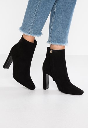 Mujer vistiendo botas de ante negras hasta el tobillo con tacones anchos y jeans azules con dobladillo deshilachado contra un fondo blanco.