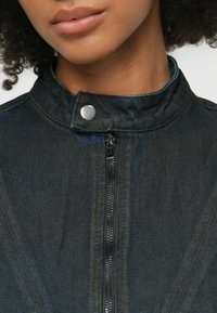 Crna traper jakna s patent zatvaračem, kopčom na dugme i teksturiranom tkaninom. Sadrži srebrne detalje i kvalitetno prošivene šavove.