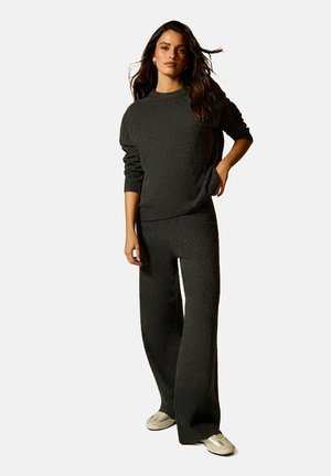 Jersey de punto gris oscuro y pantalones anchos a juego, ambos con un acabado texturizado. La modelo se sostiene con una mano en el bolsillo, vistiendo zapatillas.