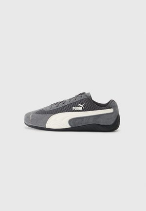 Niskoprofilowy szary sneaker Puma z białym paskiem po boku, czarną podeszwą, siateczkową i zamszową cholewką, widoczny z zewnętrznej strony na szarym tle.