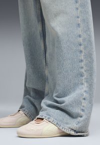 Φαρδιά τζιν ανοιχτού μπλε χρώματος φορεμένα πάνω από παπούτσια Puma σε απαλό ροζ, με λεπτομερείς ραφές και ορατό υφασμάτινο υλικό και στα δύο.