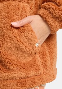 Veste en polaire couleur rouille avec une surface douce et texturée. Dotée d'une poche avant et d'une petite étiquette de marque. Les poignets ont une finition peluche.