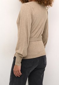 Beige knit sweater with long sleeves, fitted waist, and textured fabric. Paired with dark gray jeans, showing detail in stitching. Back view.