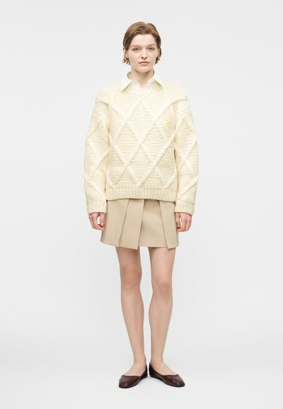 Νεαρό άτομο με κοντά μαλλιά που φοράει κρεμ πουλόβερ με πλεκτό σχέδιο, μπεζ πλισέ φούστα, λευκό πουκάμισο με γιακά και σκούρα καφέ φλατ παπούτσια, στέκεται όρθιο.