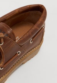 Chaussure bateau en cuir marron avec des lacets ronds, œillets en métal, semelle intermédiaire texturée beige et doublure intérieure lisse, présentant des accents de couture détaillés.