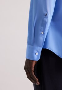 lichtblauwe blouse met lange mouwen en een gladde textuur. Heeft een puntige manchetknoopsluiting en een geborduurd detail op de mouw.