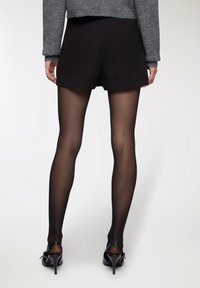 Pantaloni shorts neri abbinati a collant neri semitrasparenti e scarpe con tacco nere. Il capo presenta una texture liscia con un taglio sartoriale.