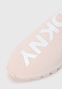 Chaussure de sport rose clair avec une tige en maille texturée, arborant un logo blanc proéminent sur le côté et une semelle en caoutchouc avec des picots antidérapants.