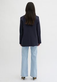 Marinblå kavaj med markerade axlar, kombinerad med ljusblå vida jeans. Bärs med svarta klackar, sedd bakifrån.