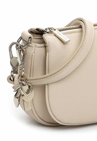 Beige Lederhandtasche mit geschwungenem Design, silbernen Beschlägen und Schlüsselanhänger-Akzenten, darunter ein Herz und Anhänger. Reißverschluss oben.